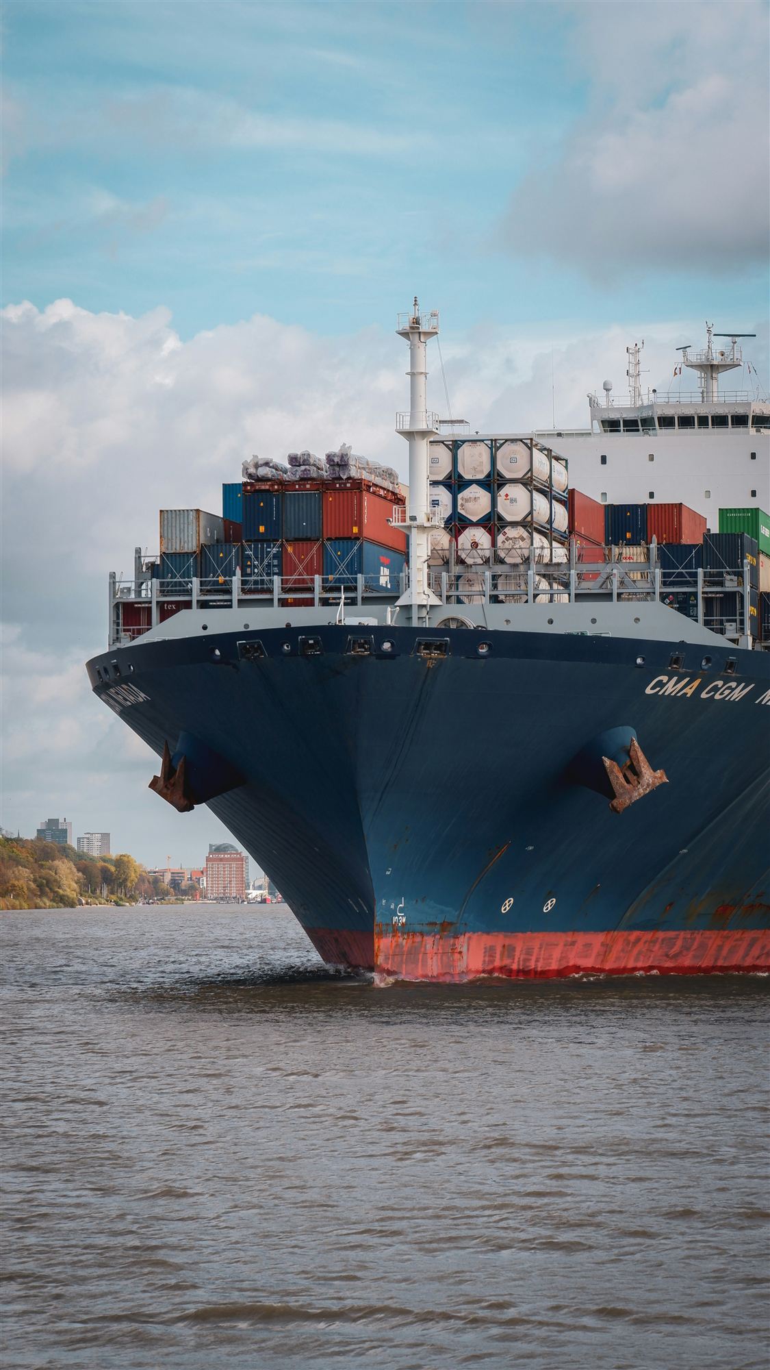 The bow of a large container ship on a river, with stacked containers and tank containers on deck.