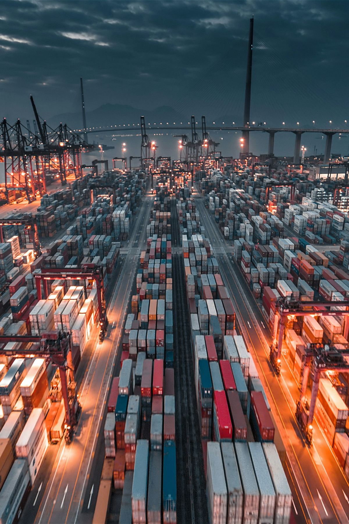 A container terminal at dusk, with quay cranes, long rows of stacked containers and a bridge in the distance.